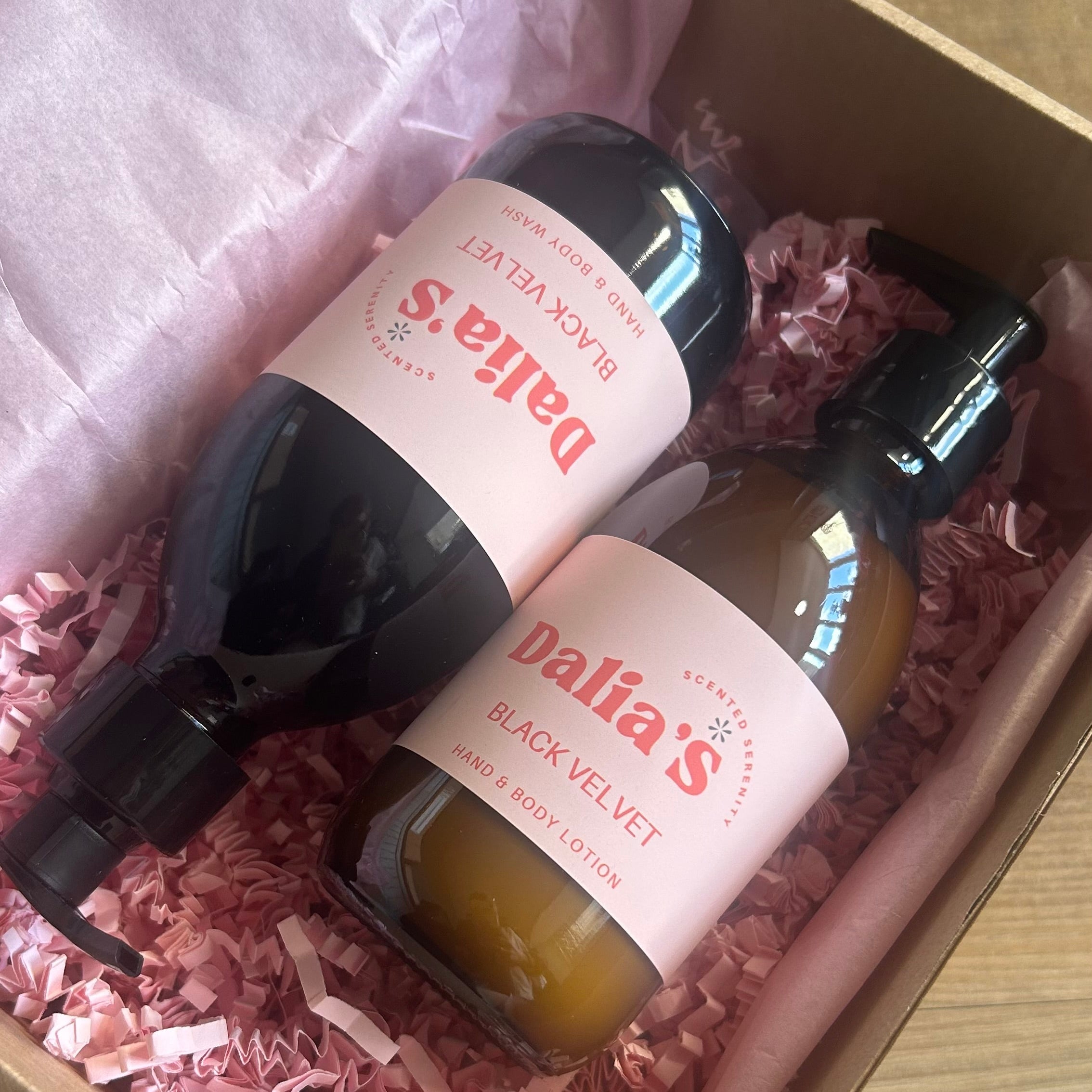 Two bottles of Dalia's products in a box with pink tissue paper on a wooden surface.