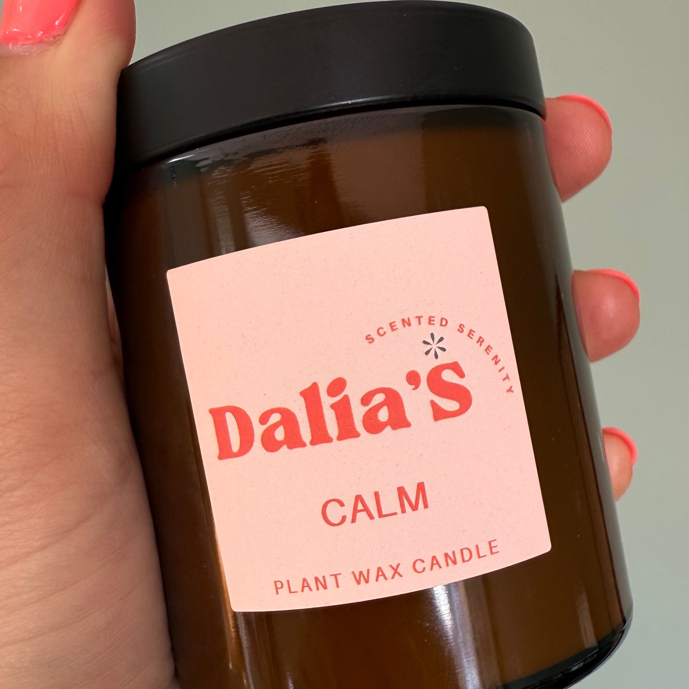 A hand holding a glass jar candle with a pink label that reads 'Dalia's Calm Plant Wax Candle'.