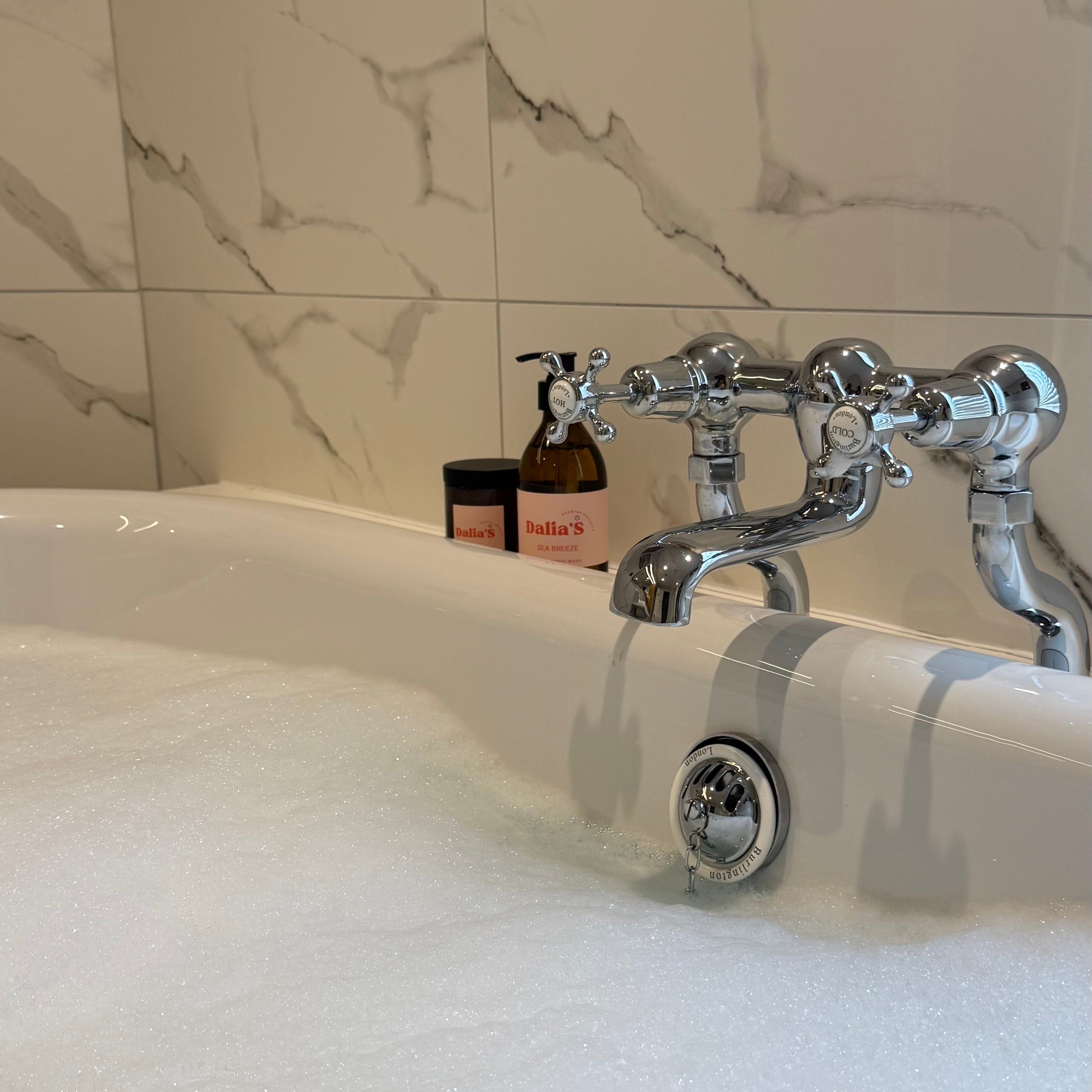 Bath tub with soaps and a bottle on the edge, against a marble tiled wall.