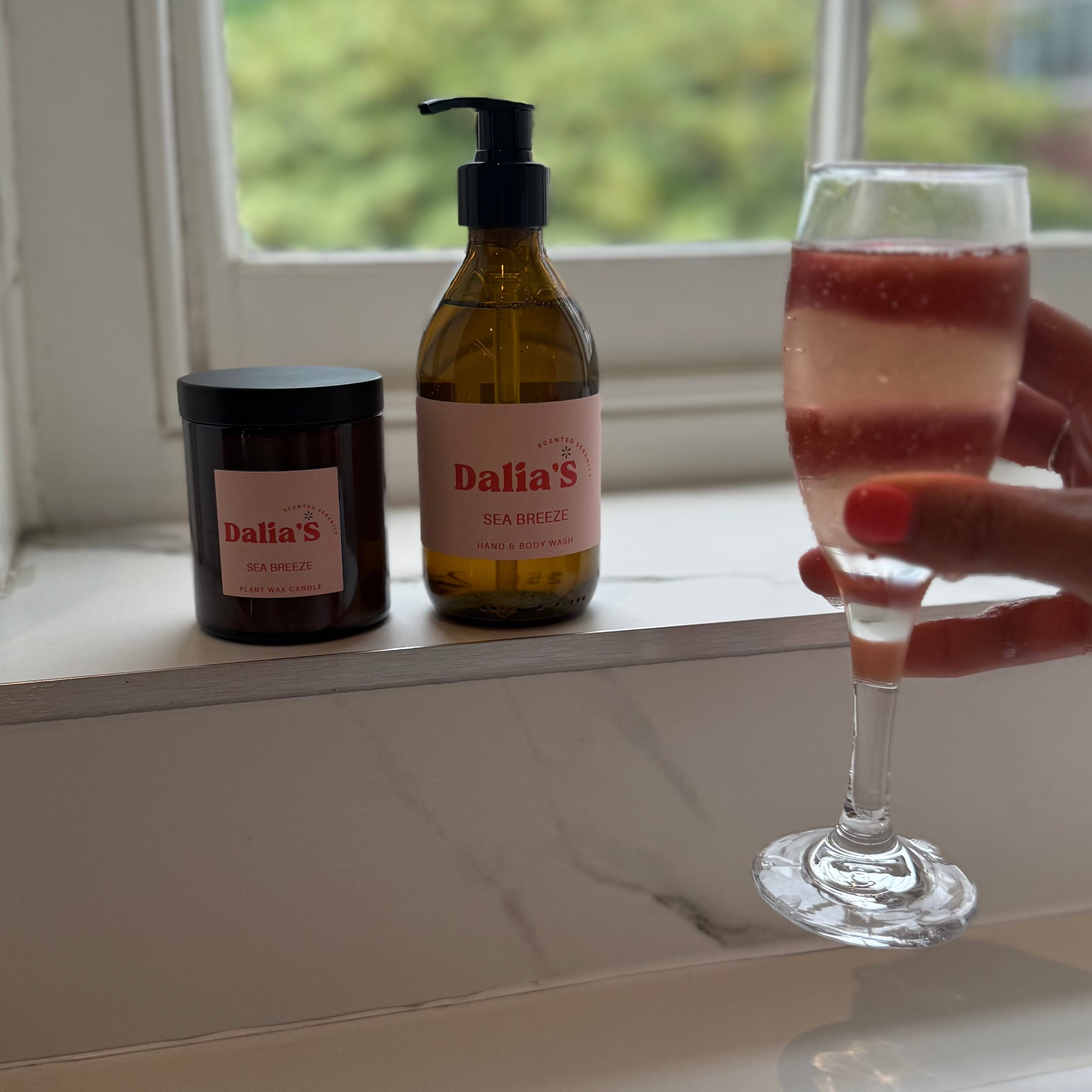 Bottle of 'Dalia's' soap, candle, and glass with a drink on a windowsill.