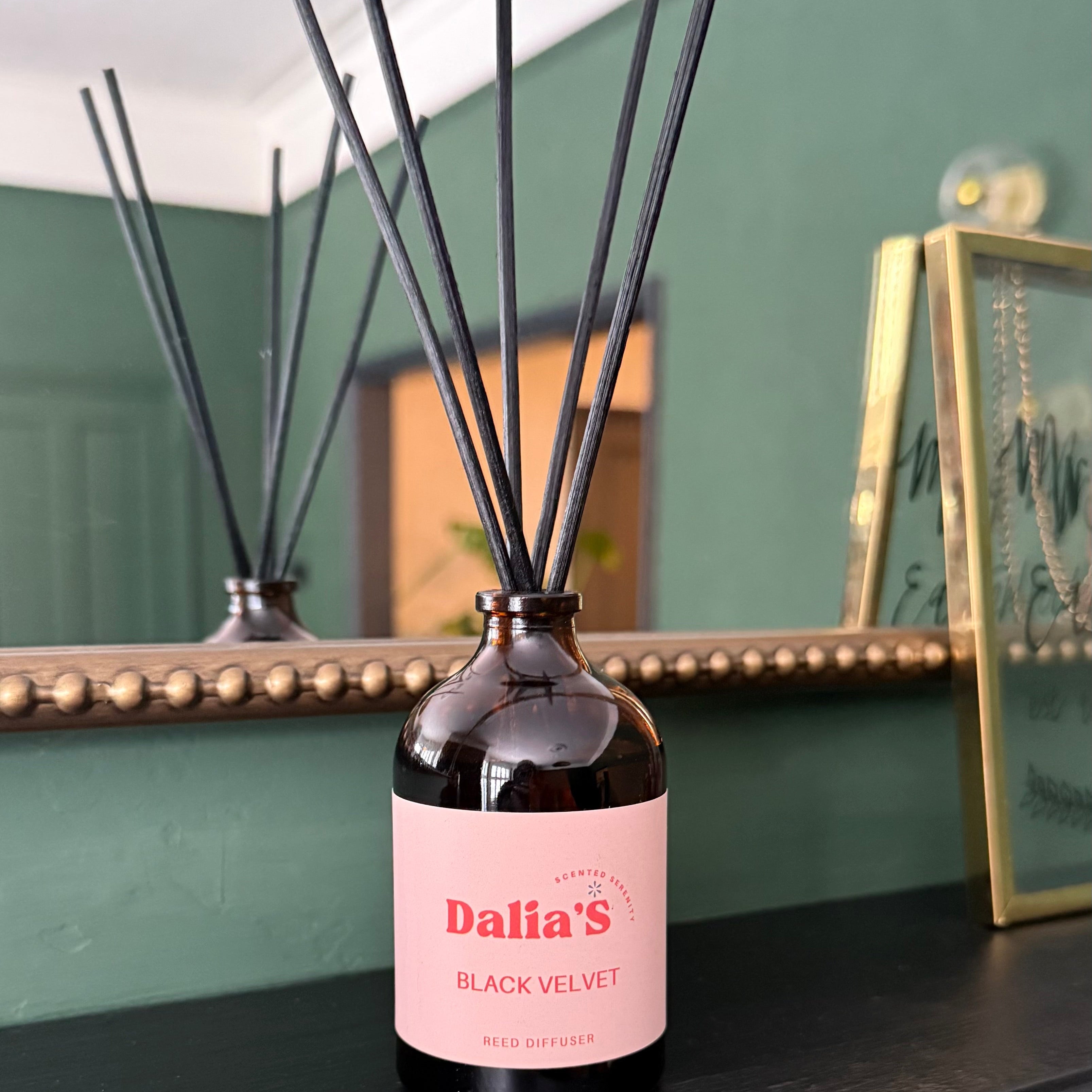 Diffuser bottle with pink label on a dark surface, with a mirror and decorative items in the background.