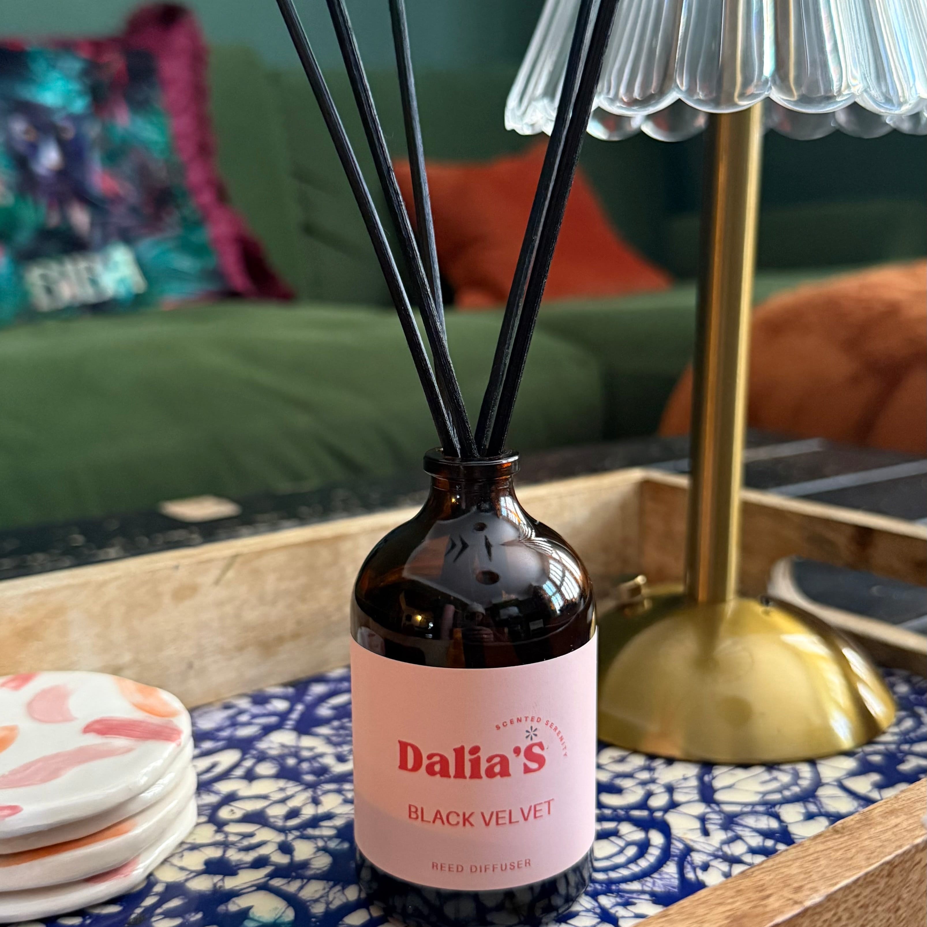 Diffuser bottle labeled 'Dalia's Black Velvet' on a decorative tray with a lamp and pillows in the background.